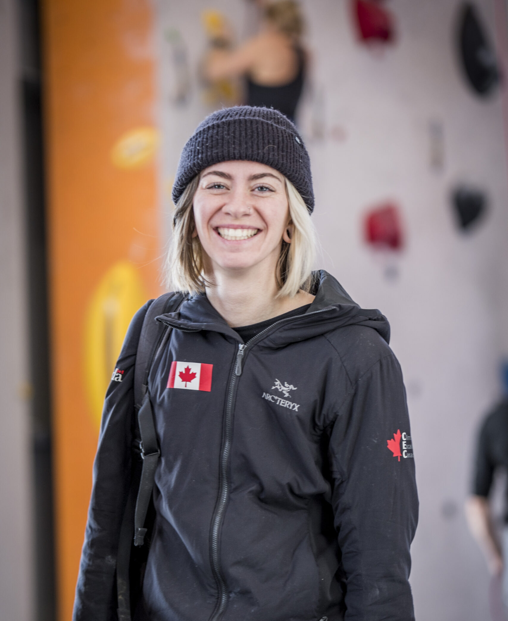 Emily wearing her black Team Canada jacket with a Canadian flag over the right side of her chest. She is shown smiling with a black toque and climbing wall in the background.